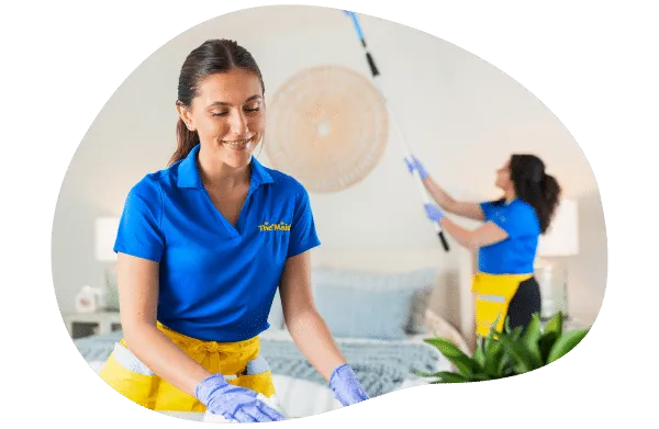 Two Maids of Scottsdale team members in blue and yellow uniforms cleaning a bedroom &mdash; one making the bed, the other dusting a light fixture.